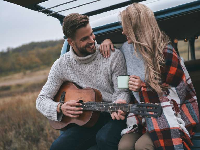 Junges Paar sitzt an der Kante eines Kofferraums und der Mann spielt Gitarre