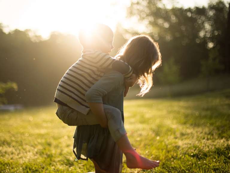 Ältere Schwester trägt jüngeren Bruder auf ihren Schultern, während sie auf einer Wiese bei Sonnenschein sind