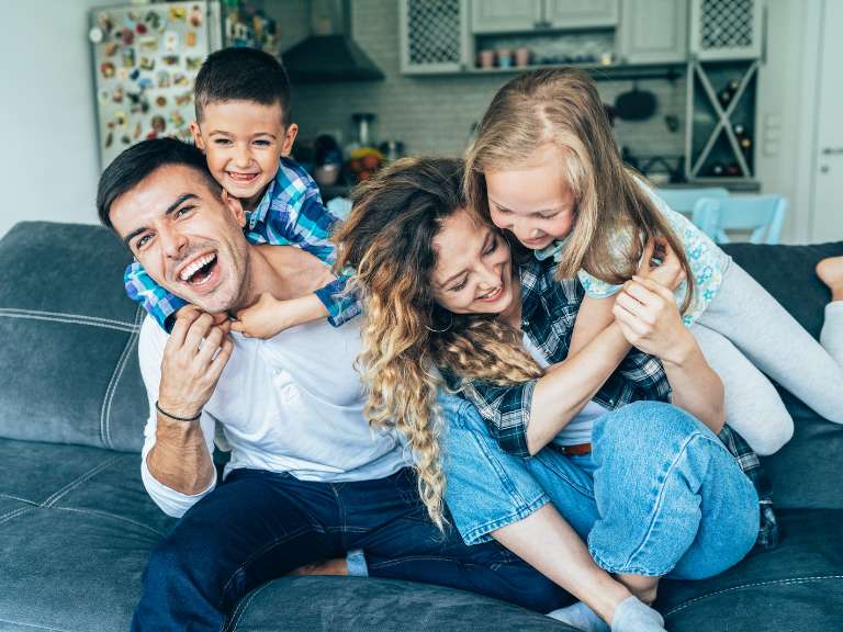 Tochter und Sohn sitzen auf dem Rücken ihrer jungen Eltern auf dem Sofa, sie haben Spaß und lachen gemeinsam