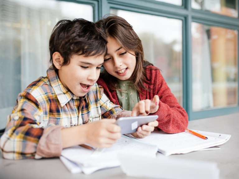 Junge und Mädchen sitzen an einem Tisch, Junge hält ein Smartphone in seiner Hand und beide schauen auf den Bildschirm des Smartphones