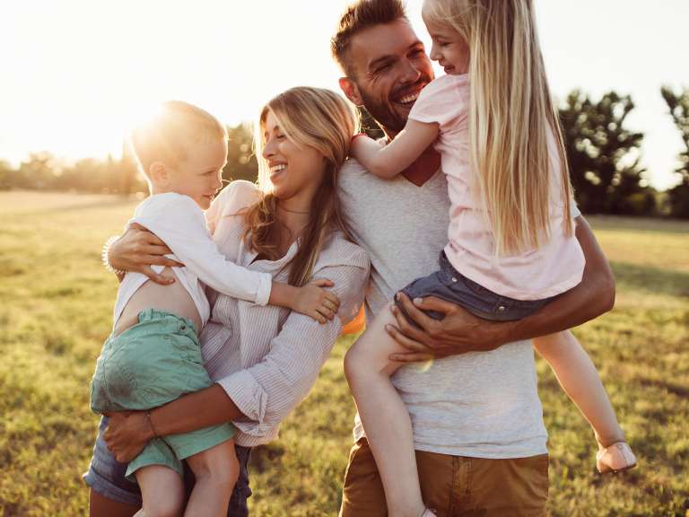 Eltern halten ihre zwei Kinder auf den Armen und lachen gemeinsam, während sie auf einer Wiese bei Sonnenschein stehen