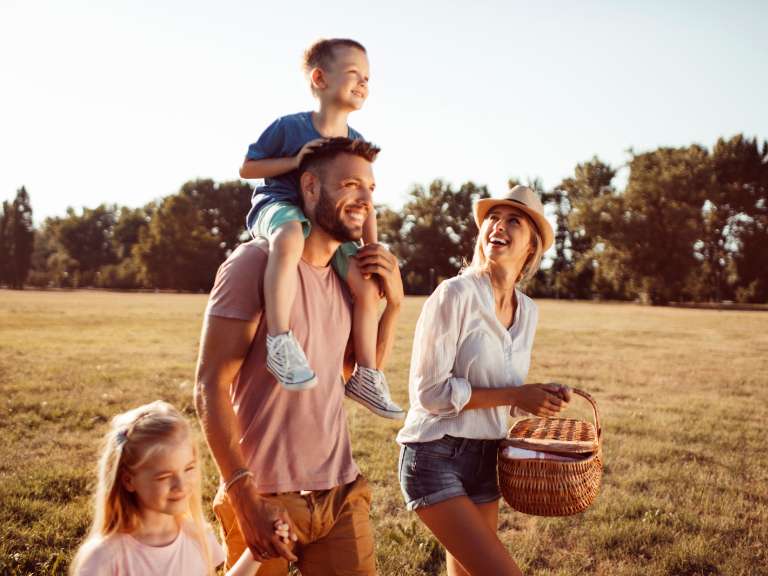 Junge Familie mit einem Sohn und einer Tochter laufen auf einer Wiese und suchen nach einem Platz für ein Picknick, Vater hält die Hand der Tochter und der Sohn sitzt auf den Schultern seines Vaters, die Mutter trägt den Picknick-Korb