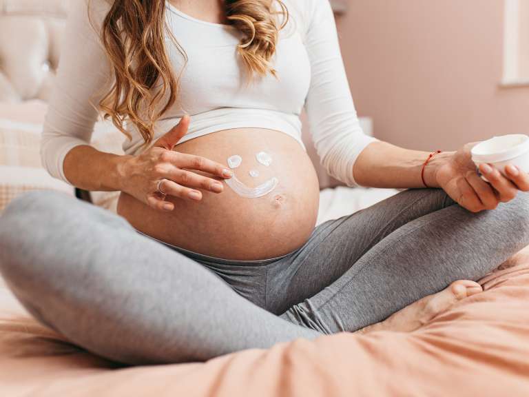 Werdende Mutter sitzt im Schneidersitz auf dem Bett und malt mit Creme einen lachenden Smiley auf ihren Babybauch
