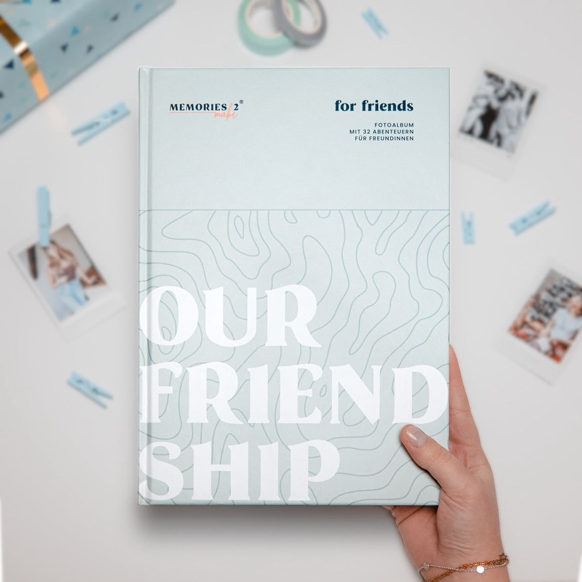 Hand hält das Challengebuch für beste Freundinnen „Our Friendship“, daneben Fotos im Polaroid-Stil und ein Geschenkpaket