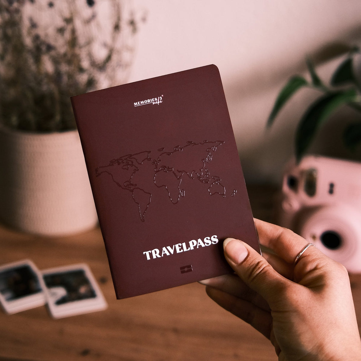 Hand hält das weinrote Reisetagebuch Travelpass im Reisepass-Design, im Hintergrund Sofortbildkamera und Fotos