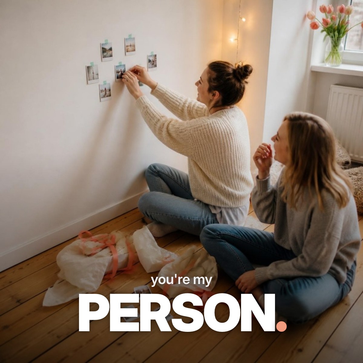 Schriftzug ‚you're my PERSON.‘ über einem Foto zweier junger Freundinnen, die auf dem Holzboden einer hellen WG ein Geschenk öffnen und kleine Fotodrucke mit Washi-Tape an die Wand kleben.