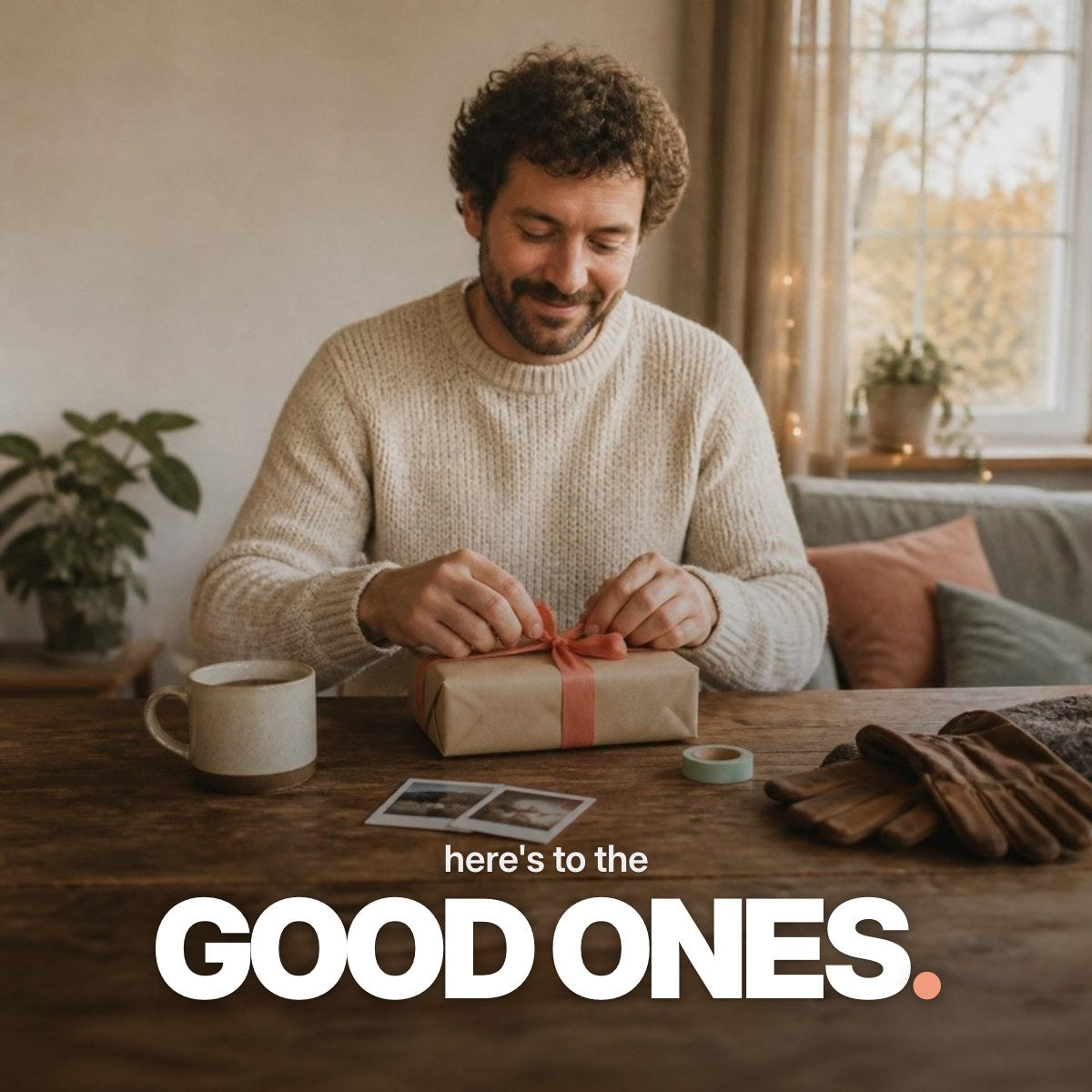 Schriftzug ‚here's to the GOOD ONES.‘ über einem warmen Foto eines Mannes im cremefarbenen Strickpullover, der an einem Holztisch neben Kaffeetasse und Sofortbildern lächelnd ein kleines Geschenk in Packpapier mit korallfarbener Schleife auspackt.
