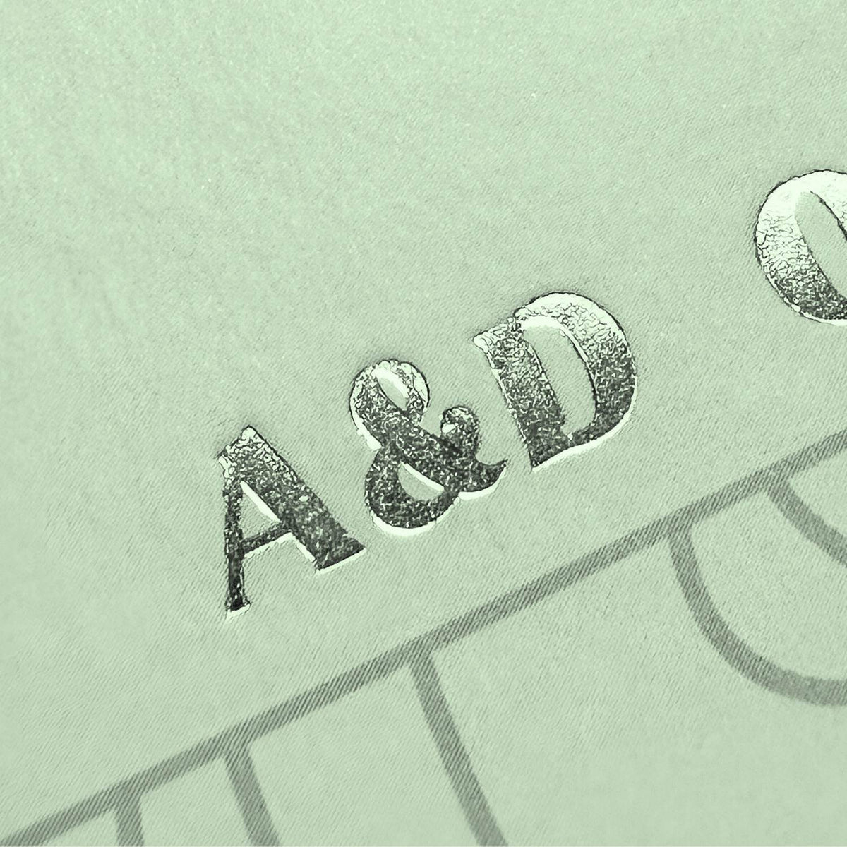 Nahaufnahme einer silbernen Folienprägung mit den Initialen „A&D“ auf einem hellgrünen Buchcover