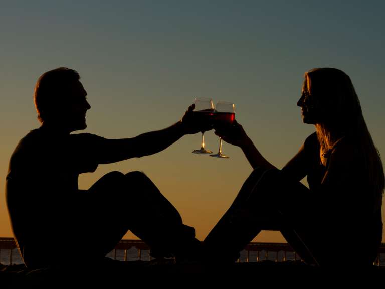 Junges Paar sitzt sich gegenüber auf einer Mauer bei Sonnenuntergang und stoßt mit einem Glas Wein an