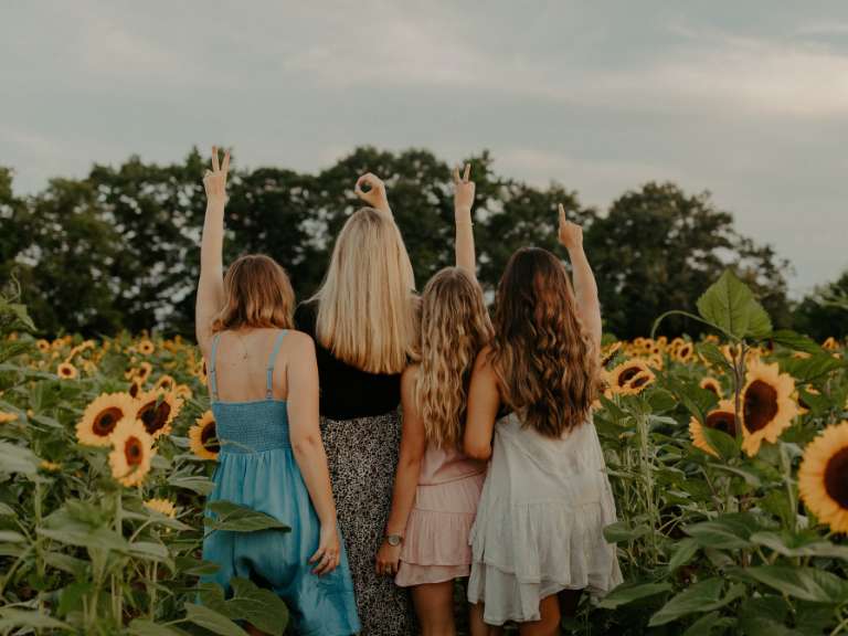Vier Freundinnen stehen nebeneinander in einem Feld voller Sonnenblumen und strecken die Arme nach oben