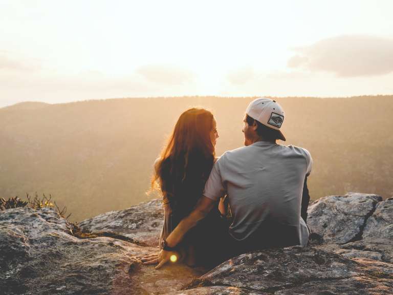 Junges Paar sitzt auf einem Fels, umarmt sich und beobachtet den Sonnenaufgang