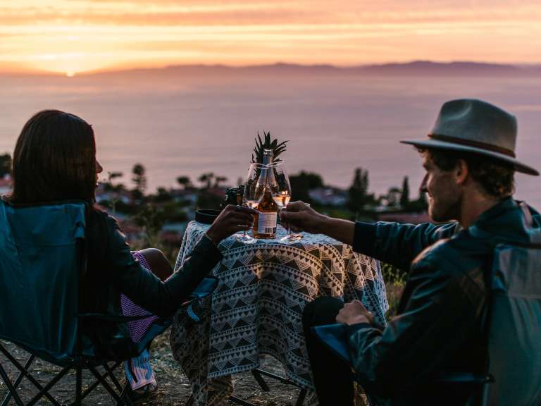Mann und Frau sitzen bei Sonnenuntergang nebeneinander an einem Tisch und stoßen mit einem Glas Sekt an