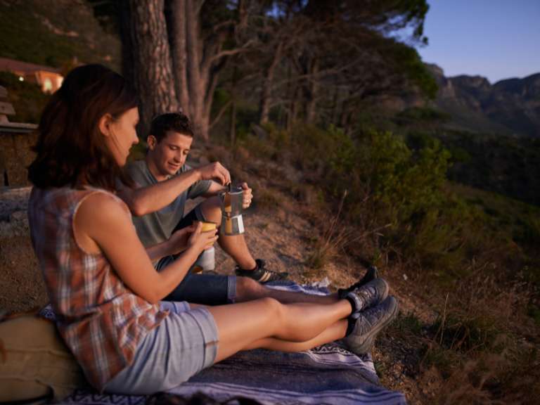 Mann und Frau sitzen gemeinsam bei Sonnenschein an einem Berg und schenken Kaffee in eine Tasse