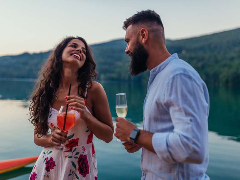 Junges Paar steht mit einem Weinglas vor einem See und lacht gemeinsam