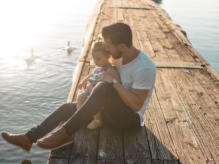 Vater und Tochter sitzen auf einem Steg an einem See und Tochter sitzt auf dem Schoß des Vaters
