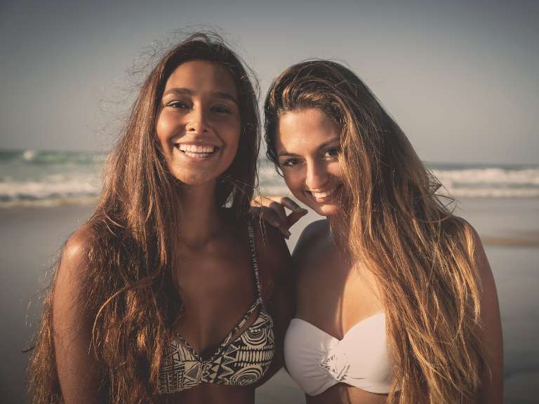 Zwei Freundinnen sind am Strand am Meer im Bikini und lächeln in die Kamera