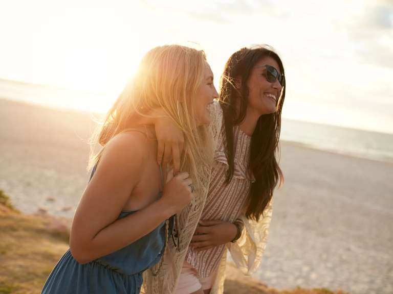 Zwei Freundinnen sind bei Sonnenschein am Meer und laufen Arm in Arm am Strand entlang