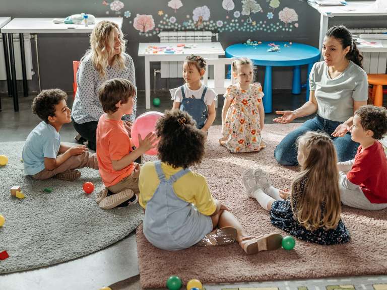 Erzieherinnen und Kinder sitzen in einem Kreis auf dem Boden in einem Kindergarten und sprechen miteinander