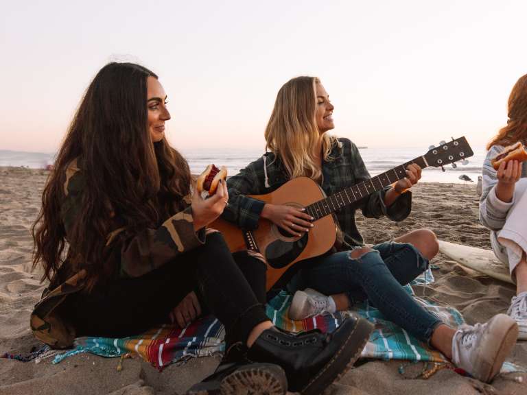 Zwei junge Frauen sitzen bei einem Lagerfeuer am Strand, eine davon spielt auf der Gitarre