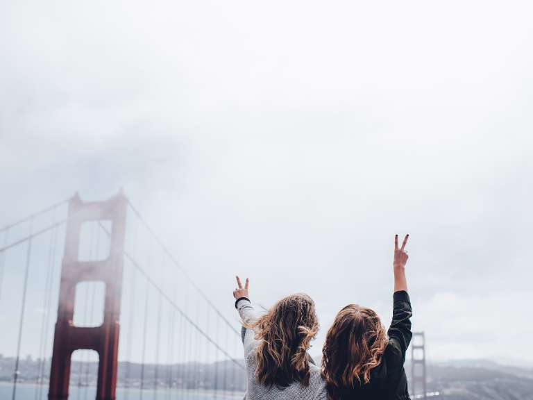 Zwei junge Frauen sind gemeinsam im Urlaub, stehen an einer Brücke und strecken ihre Arme nach oben