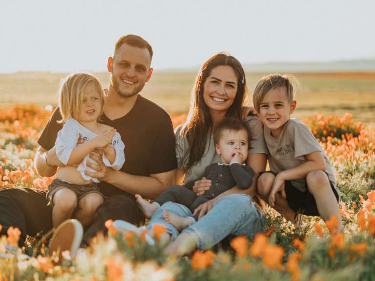 Junge Familie mit drei Kindern sitzt bei Sonnenschein auf einer Blumenwiese und lächelt in die Kamera