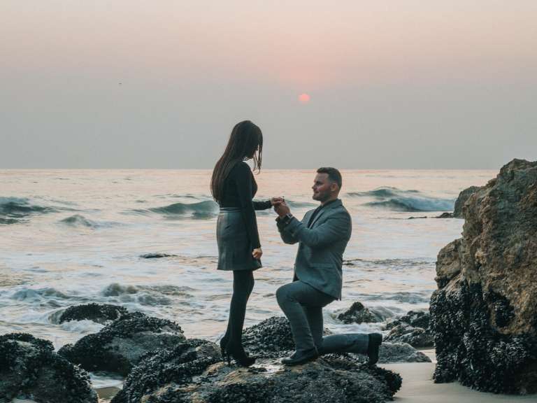 Mann kniet vor Frau auf den Boden auf einem Fels am Meer und macht ihr einen Heiratsantrag