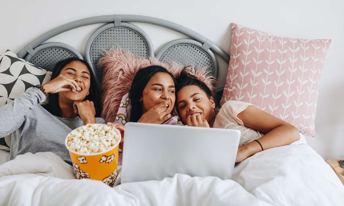 Drei Freundinnen unter einer weißen Decke essen Popcorn und schauen lachend zusammen einen Film auf dem Laptop