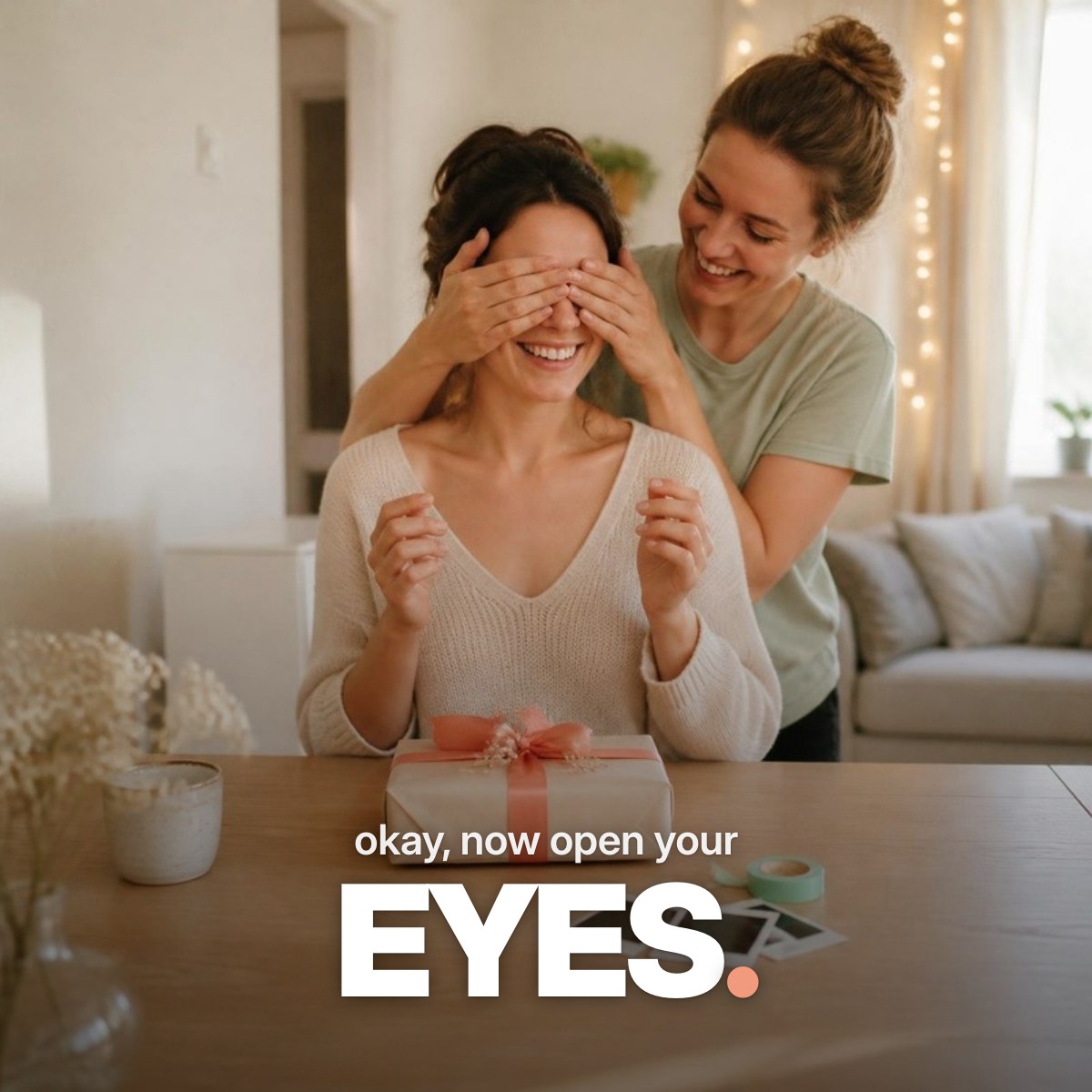 Schriftzug ‚okay, now open your EYES.‘ über einem Foto, auf dem eine junge Frau ihrer lachenden besten Freundin in einem hellen Wohnzimmer von hinten die Augen zuhält, vor ihnen ein Geschenk mit korallfarbener Schleife und Sofortbilder.