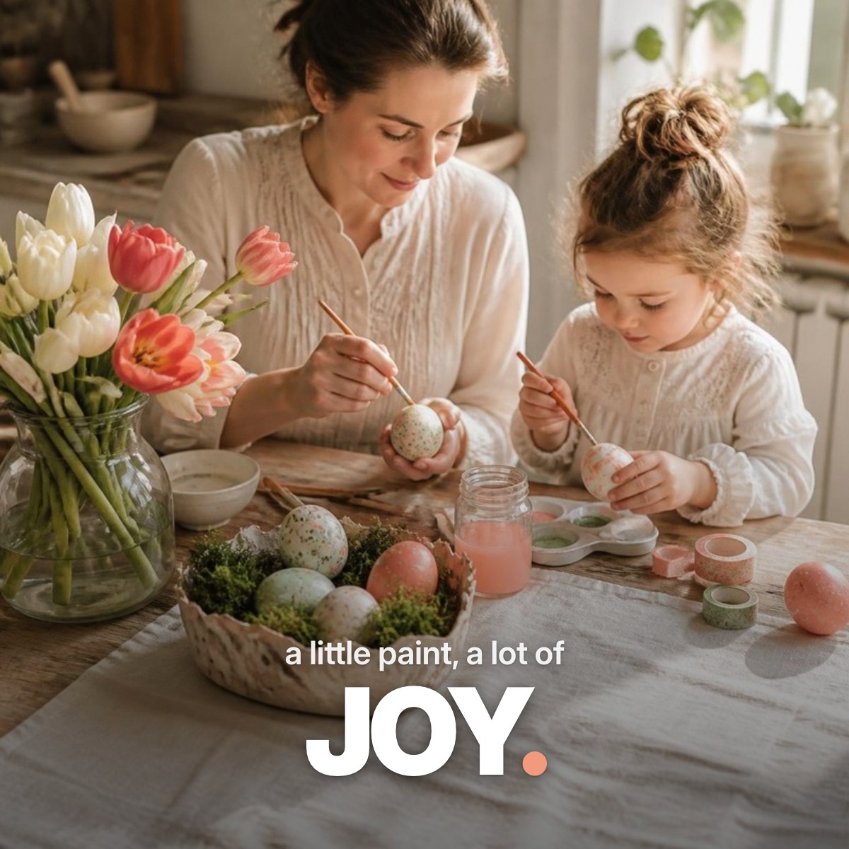 Schriftzug ‚a little paint, a lot of JOY.‘ über einem Foto, auf dem die Hände einer Frau und eines Kindes an einem hellen Küchentisch mit Tulpen und Osterkörbchen Ostereier in Pastellfarben bemalen.