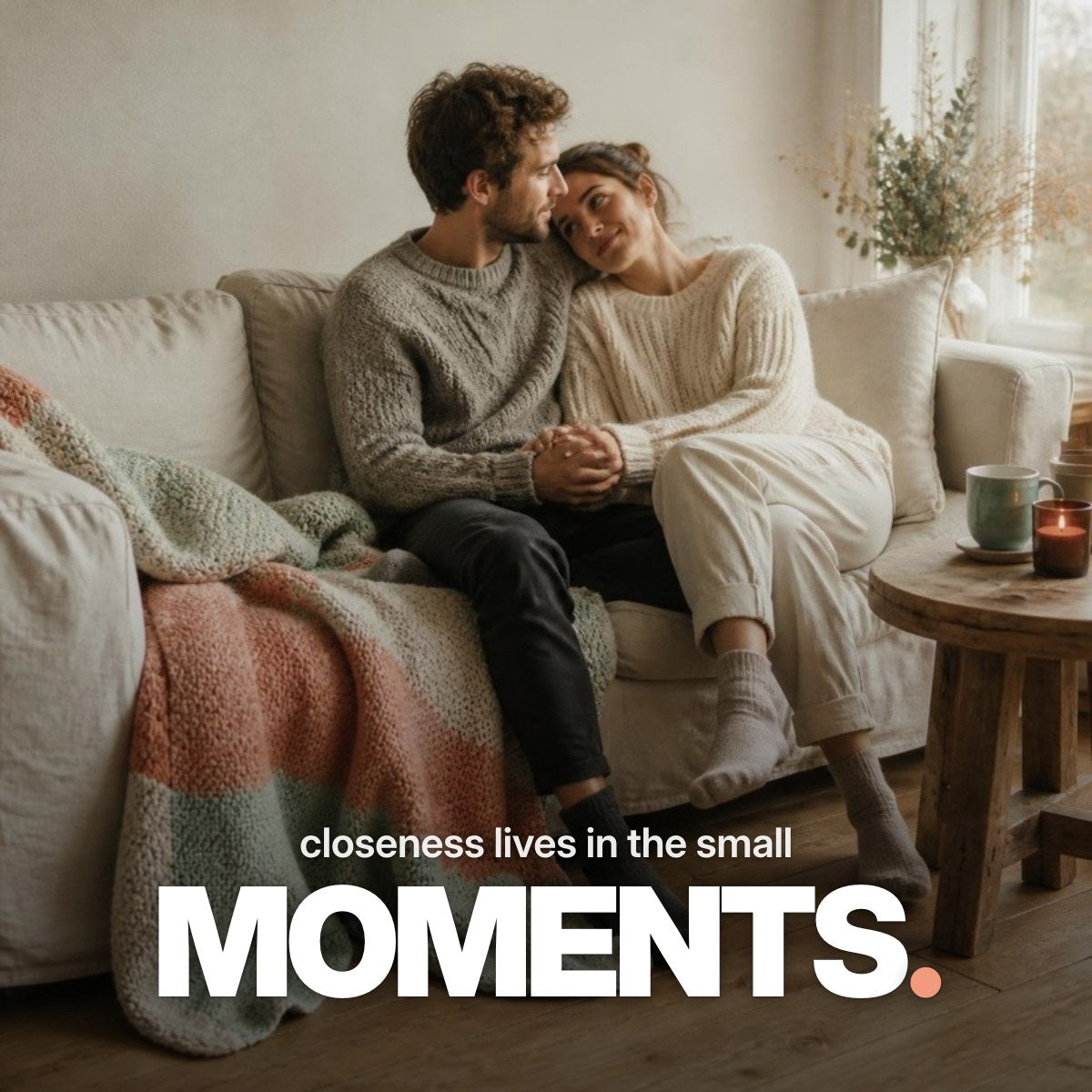 Schriftzug ‚closeness lives in the small MOMENTS.‘ über einem Foto eines Paares in Strickpullovern, das eng beieinander auf einem Sofa sitzt, Hand in Hand, mit Teetassen und Kerze im warmen Tageslicht.