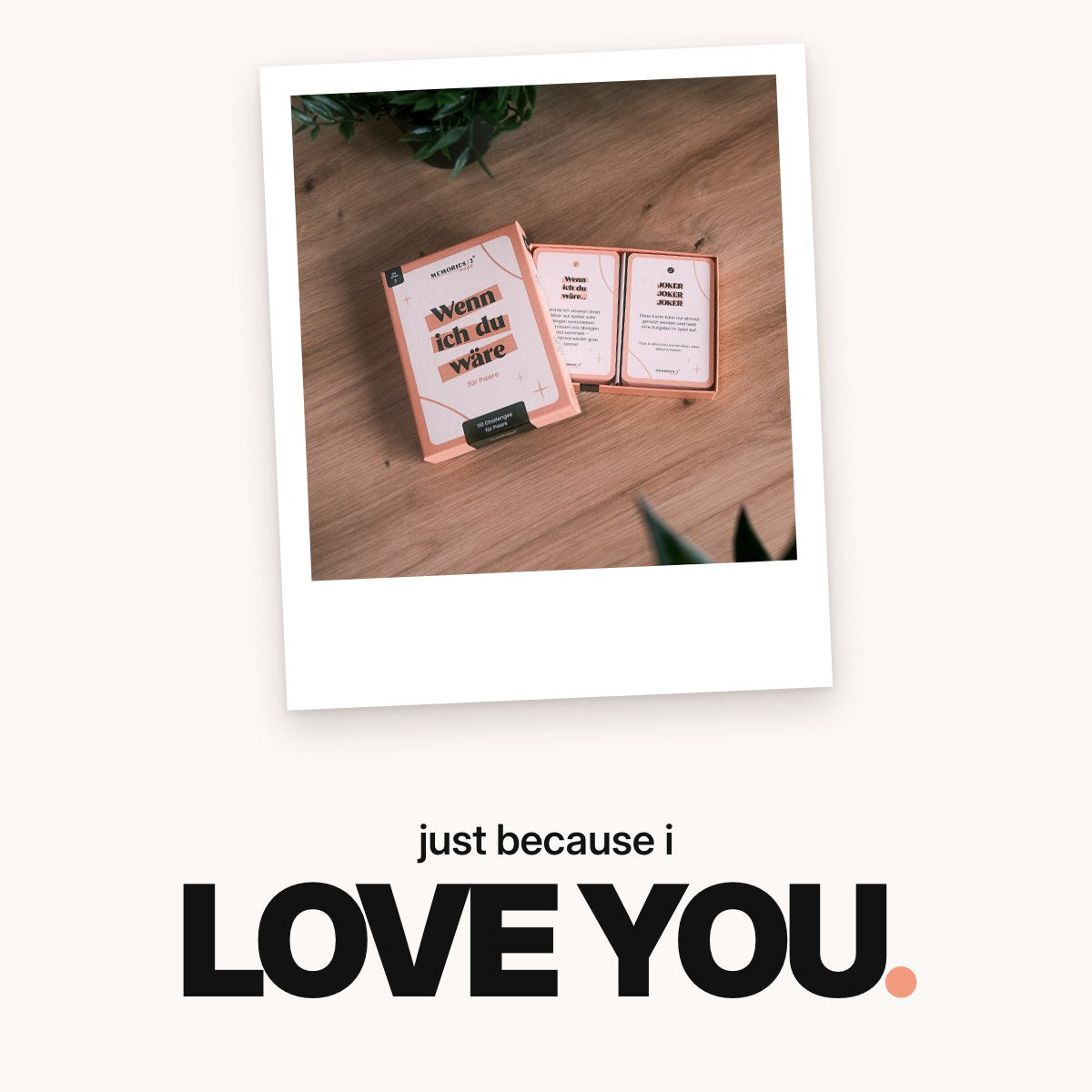 Schriftzug ‚just because i LOVE YOU.‘ über der geöffneten Schachtel des Paar-Kartenspiels „Wenn ich du wäre“ mit Aufgaben- und Jokerkarte auf einem Holztisch zwischen Grünpflanzen.